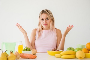 Mujer con expresión de asombro o frustración, sentada frente a una mesa con frutas y verduras, representando la confusión o desánimo al no ver resultados rápidos pese a comer saludable.