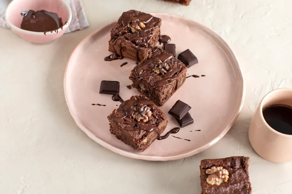 varios trozos de Brownie encima de un plato junto con tozos de chocolate negro y cerquita una taza con café negro. Bizcocho hecho con harinas sin gluten y chocolate negro. Sin problemas de intolerancia