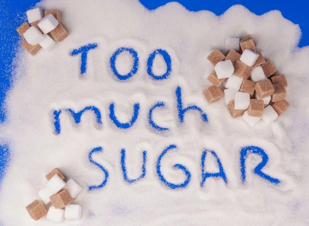 Imagen con azúcar molida y azucarillos escribiendo sobre el azúcar en una encimera azul que es "demasiado azúcar", no hay que abusar en una dieta saludable. >No apto para dulces saludables